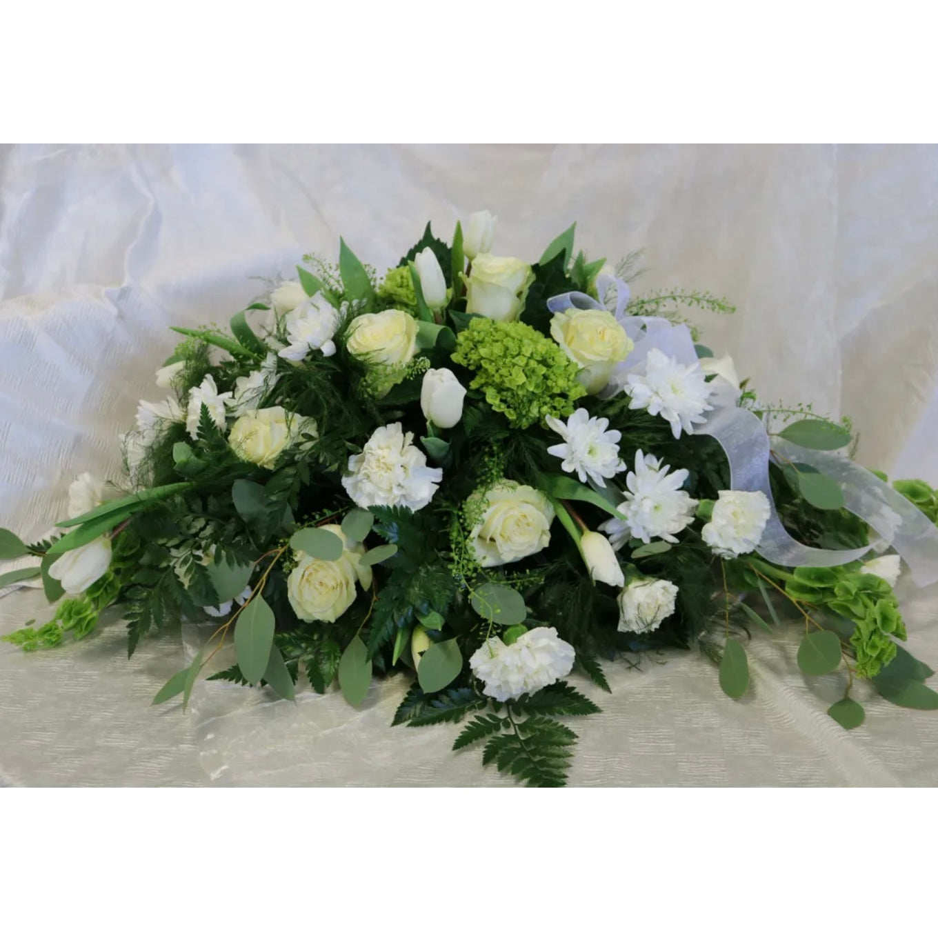 The Serenity - Casket Spray by Posies Flowers & Fashion is an exquisite floral arrangement featuring a variety of white roses, tulips, and carnations interspersed with green leaves and ferns, elegantly displayed in a sophisticated container against a soft, white fabric backdrop. Be sure to check for availability so you don't miss out on this stunning display.