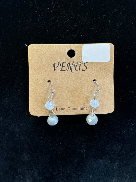 A pair of stylish Grey Earrings showcased on a brown card featuring "VENUS" text. The earrings have an elegant grey beaded design, each adorned with a small translucent light blue bead and a larger faceted light blue bead, dangling from silver hooks. The background is black fabric, and the card notes "Lead Complaint," branded by Posies Flowers & Fashion Inc.