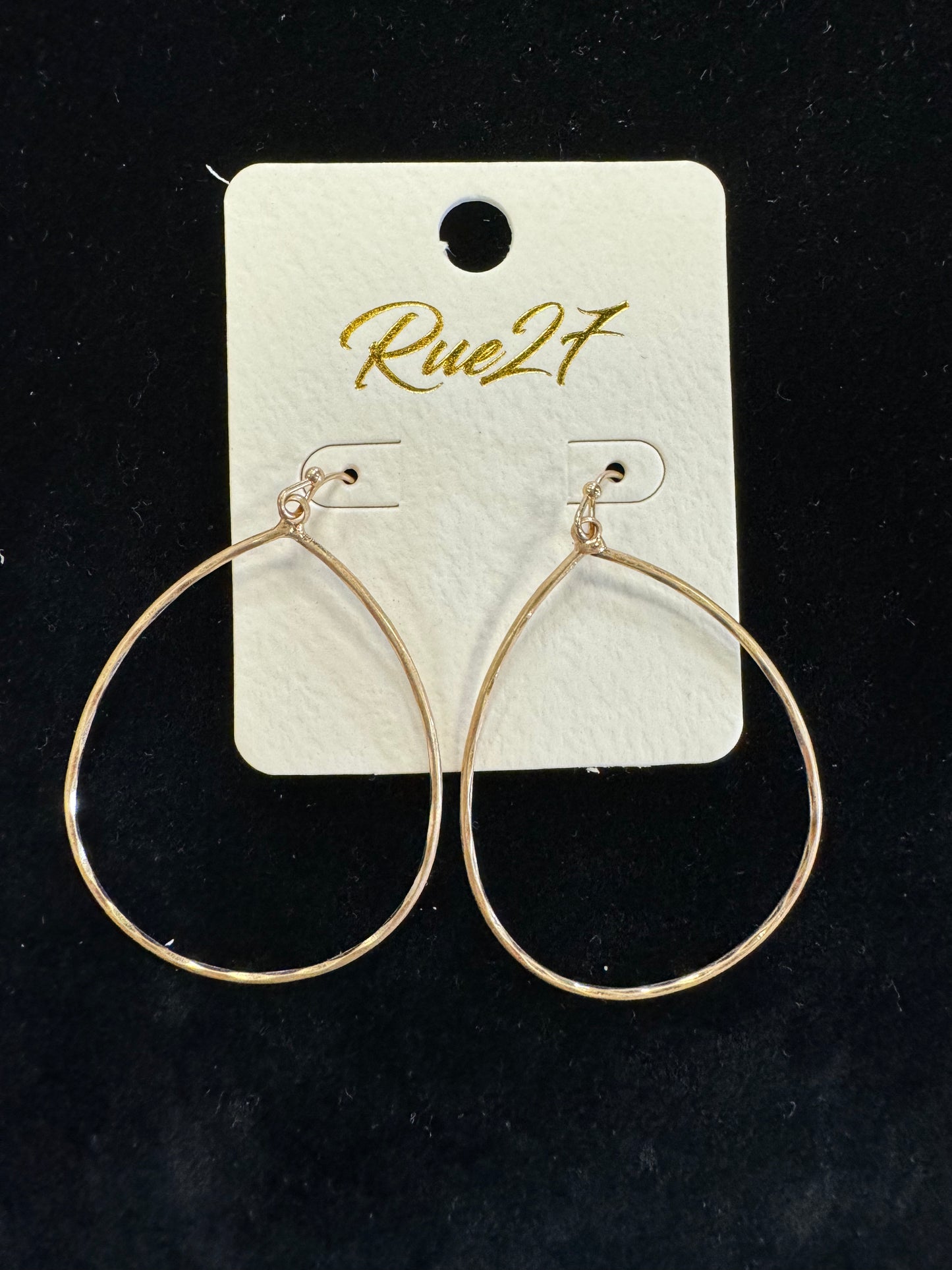 A pair of thin, gold-colored teardrop-shaped hoop earrings are displayed on a white card with the brand name "Posies Flowers & Fashion Inc." in gold cursive writing. The card is set against a plain black background. This elegant addition to any jewelry collection radiates timeless charm.