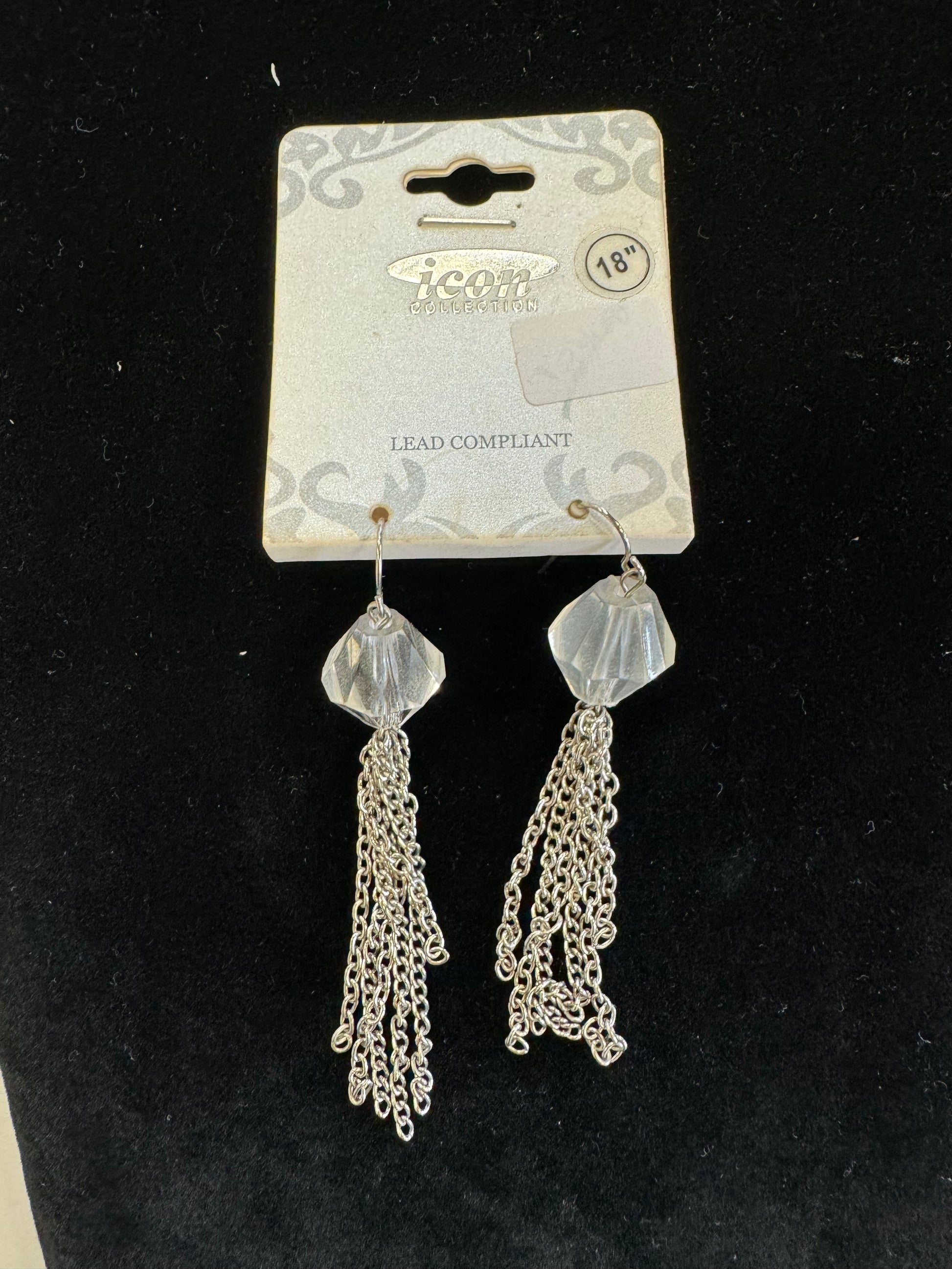 A pair of iconic earrings from Posies Flowers & Fashion Inc. is displayed against a black background. Each earring features a large, faceted translucent bead attached to multiple silver chain tassels. The chic dangle details are mounted on a white card labeled "Iconic Earrings" and "LEAD COMPLIANT" with an age warning of 18+.