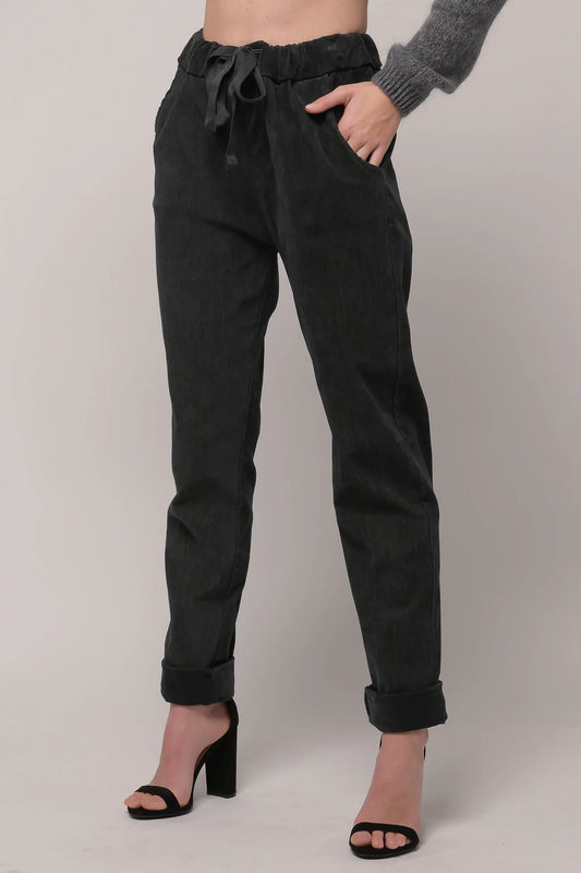 Person standing against a plain background, wearing the EKRU - BRUSHED CORD PANT - ANTRACITE from EKRU europe, which features high-waisted design, black color with a drawstring waist, pockets, and rolled cuffs. They are also wearing black heeled sandals and a gray long sleeve sweater from LINEN LUV.