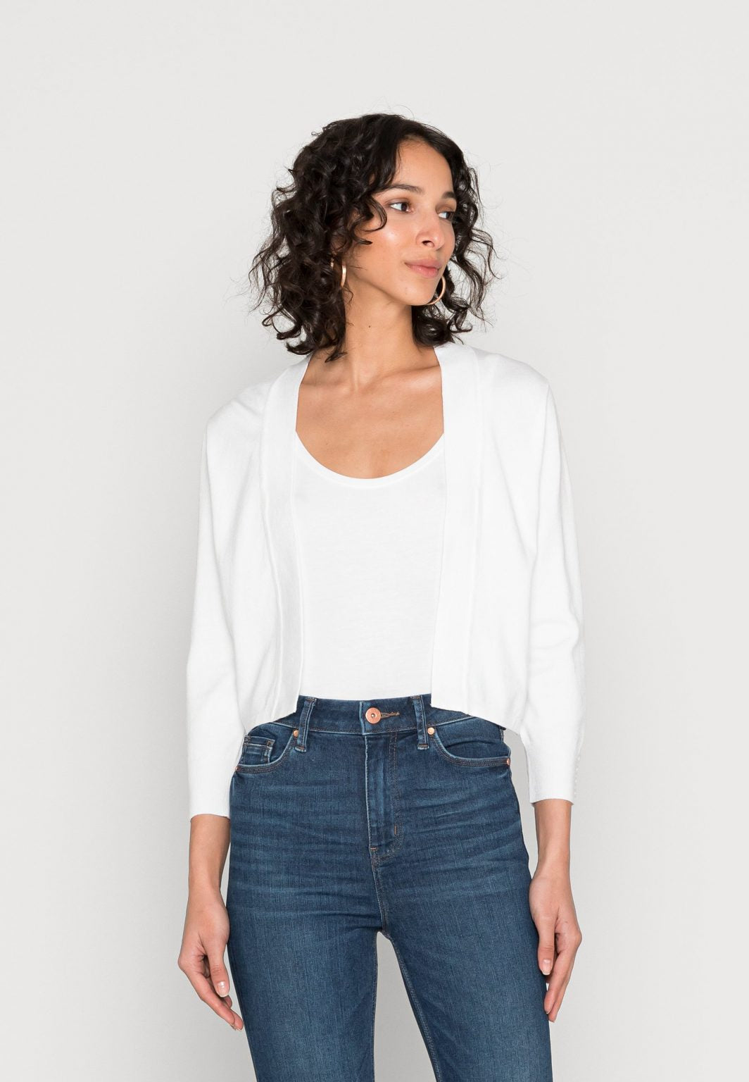 A woman with curly hair stands against a plain white background. She is wearing a white top, the White Cardigan SC-DOLLIE 662 by SoyaConcept, blue jeans, white sandals, and is holding a beige handbag. She poses with one hand on her hip, exuding confidence.