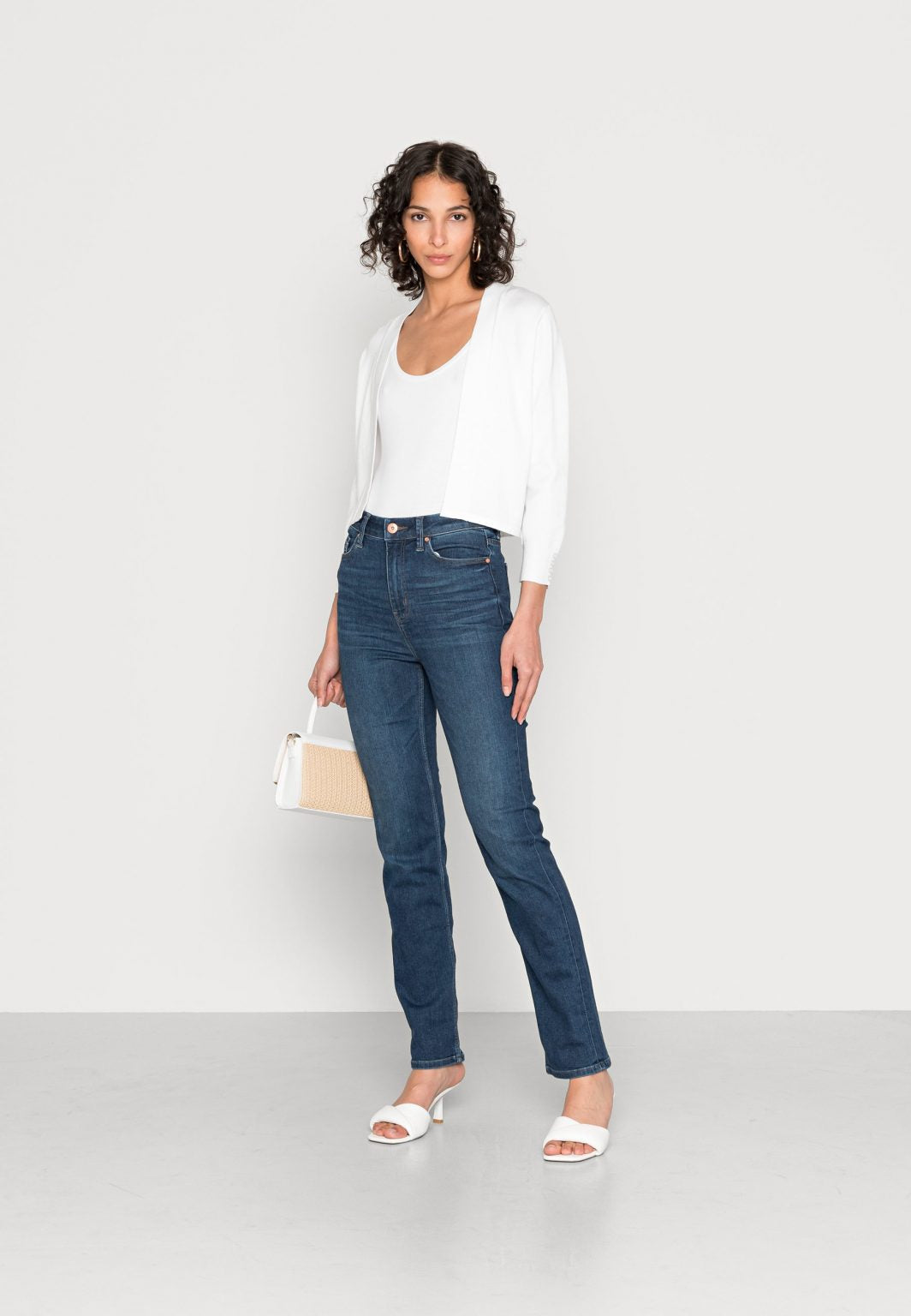 A woman with curly hair stands against a plain white background. She is wearing a white top, the White Cardigan SC-DOLLIE 662 by SoyaConcept, blue jeans, white sandals, and is holding a beige handbag. She poses with one hand on her hip, exuding confidence.