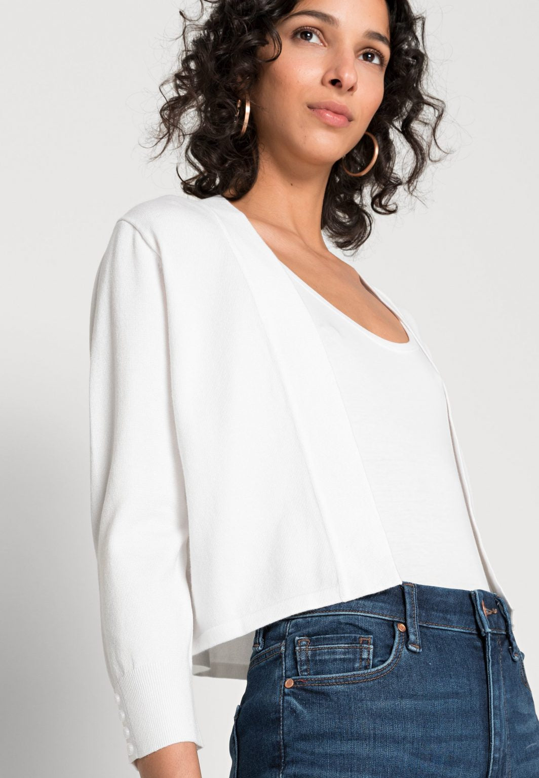 A woman with curly hair stands against a plain white background. She is wearing a white top, the White Cardigan SC-DOLLIE 662 by SoyaConcept, blue jeans, white sandals, and is holding a beige handbag. She poses with one hand on her hip, exuding confidence.