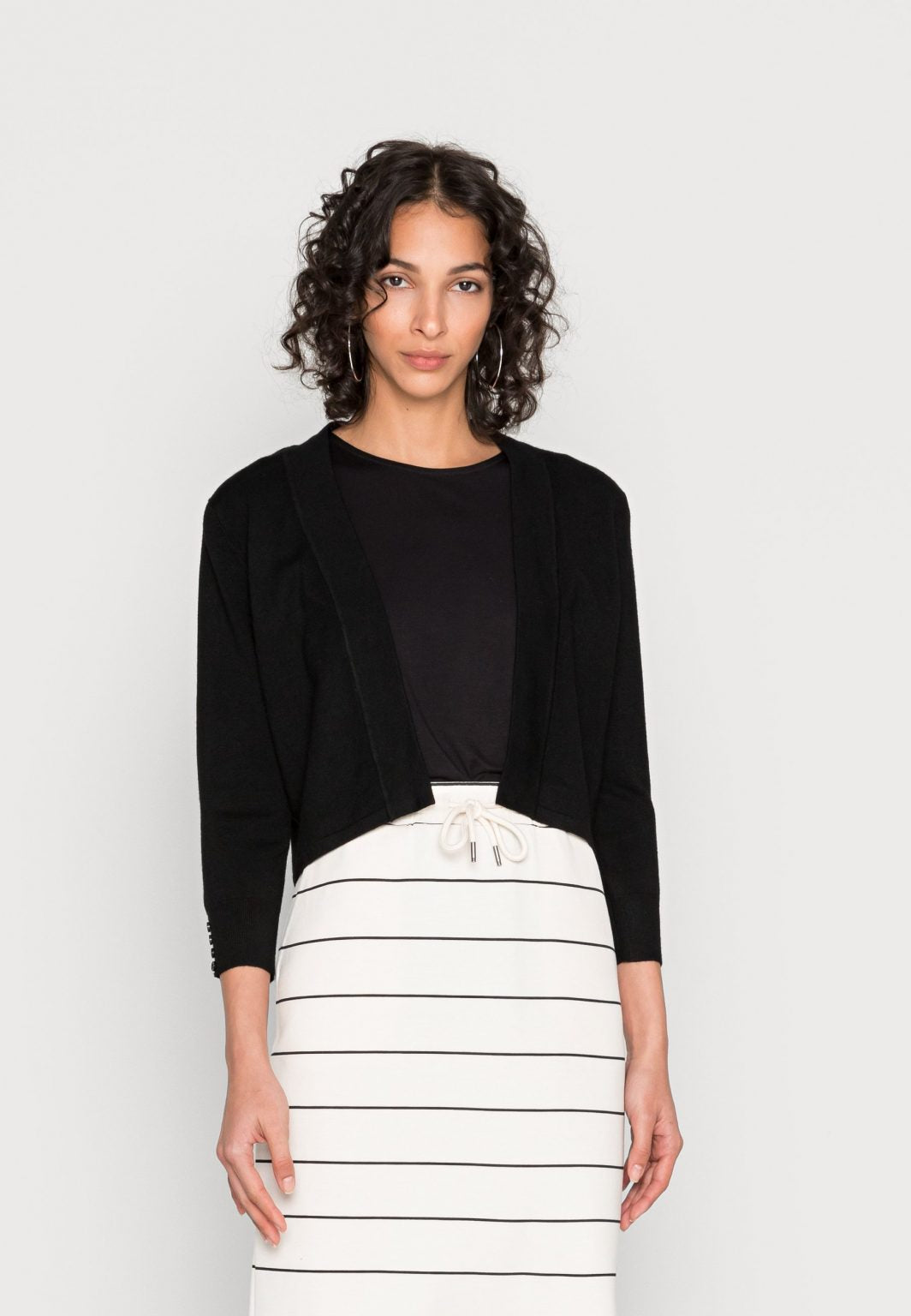 A person with curly hair is wearing a black long-sleeve Black Cardigan - SC-DOLLIE 662 - Soyaconcept over a black top and a white skirt with black horizontal stripes. The individual stands against a plain white background, looking directly at the camera.