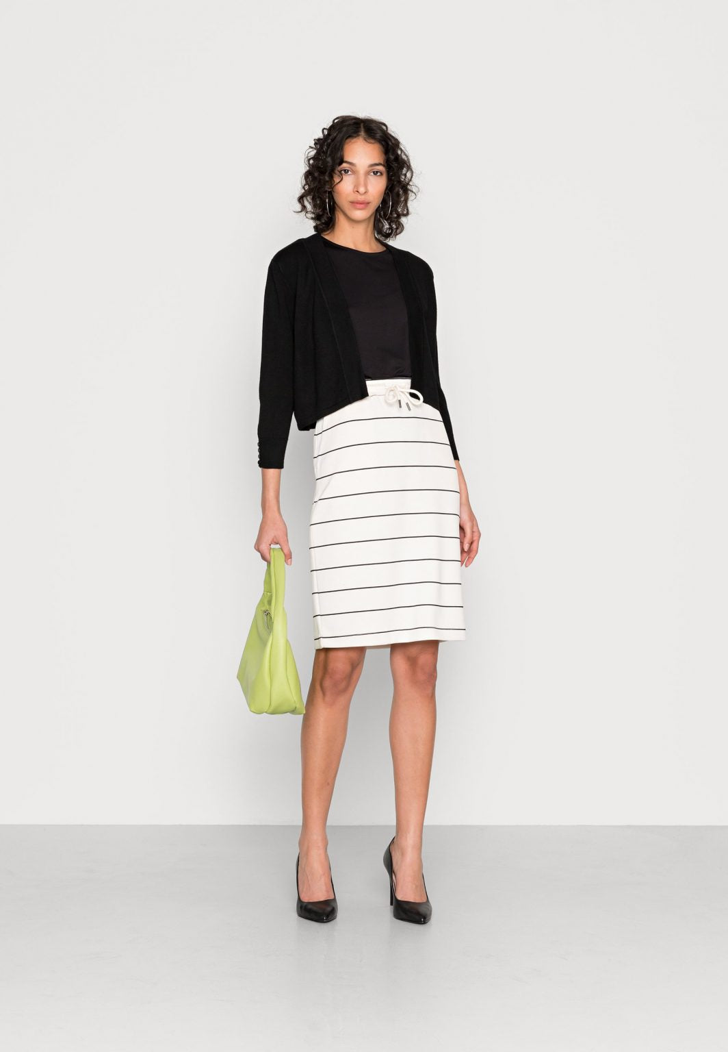 A person with curly hair is wearing a black long-sleeve Black Cardigan - SC-DOLLIE 662 - Soyaconcept over a black top and a white skirt with black horizontal stripes. The individual stands against a plain white background, looking directly at the camera.