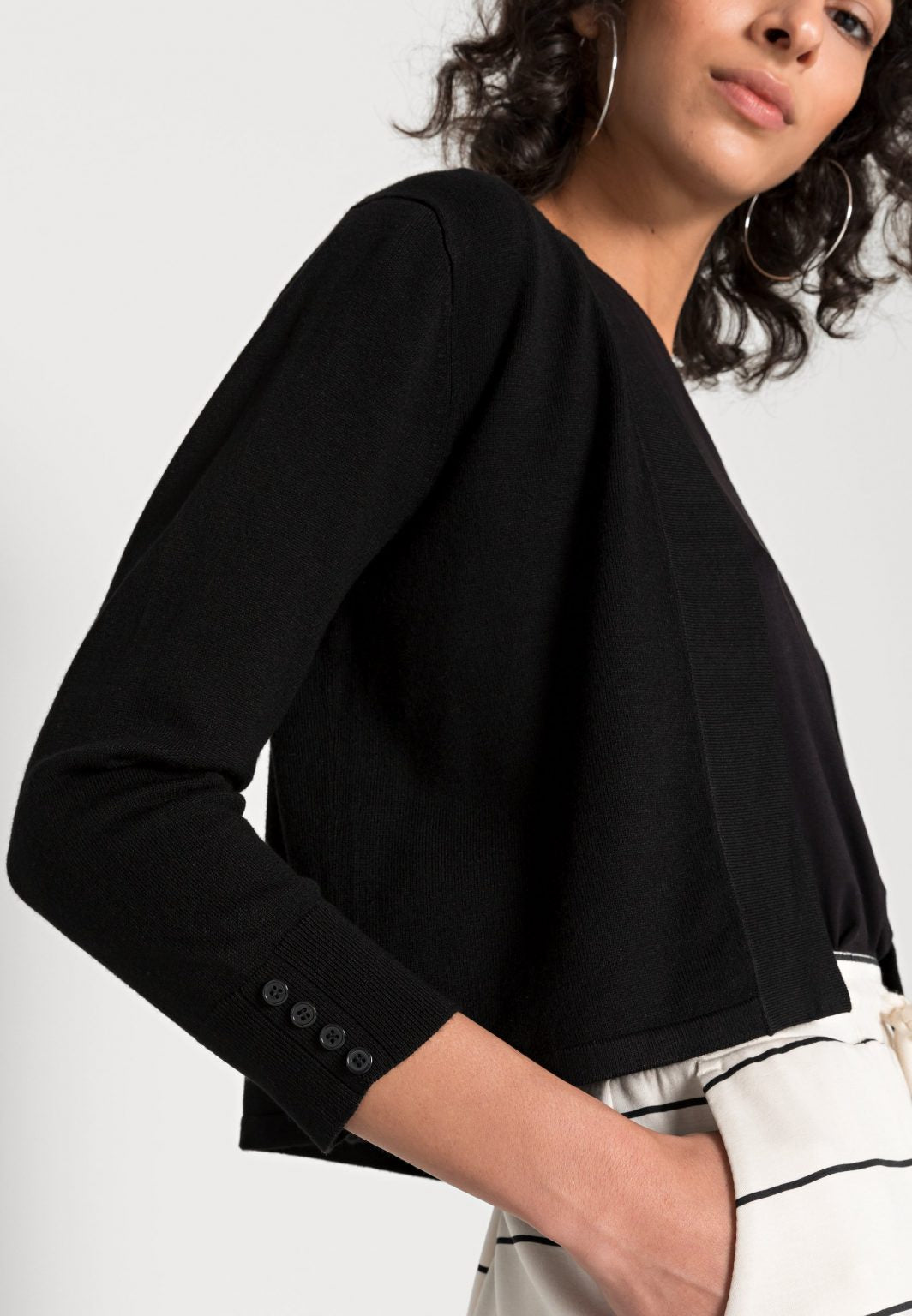 A person with curly hair is wearing a black long-sleeve Black Cardigan - SC-DOLLIE 662 - Soyaconcept over a black top and a white skirt with black horizontal stripes. The individual stands against a plain white background, looking directly at the camera.