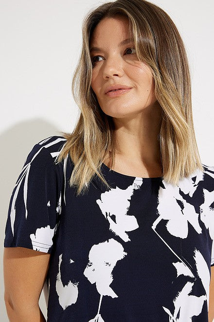 A woman with shoulder-length blonde hair, dressed in a navy and white Floral Print Tunic Style 231262 by Joseph Ribkoff, is looking up to her right. She pairs this stylish top with sleek pants. The background is plain white.