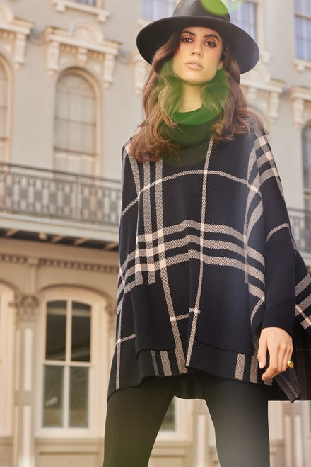 A woman stands confidently in front of an ornate building with tall windows. She is wearing a large black hat and Joseph Ribkoff's stylish Plaid Tunic Style 233965 in brown and beige, featuring a cozy cowl neck, over black pants—perfect for chilly days. Sunlight filters into the frame, creating a soft glow.