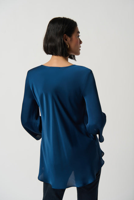 A person with short black hair is wearing a Royal Sapphire Draped Satin Blouse (Style 234082) from Joseph Ribkoff, featuring three-quarter sleeves and a tiered hem that adds an elegant touch. They are also wearing black pants. They are standing against a plain white background, gazing at the camera with arms loosely crossed.