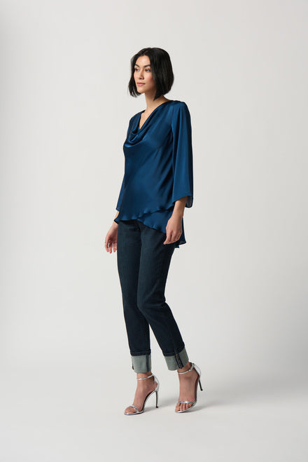 A person with short black hair is wearing a Royal Sapphire Draped Satin Blouse (Style 234082) from Joseph Ribkoff, featuring three-quarter sleeves and a tiered hem that adds an elegant touch. They are also wearing black pants. They are standing against a plain white background, gazing at the camera with arms loosely crossed.