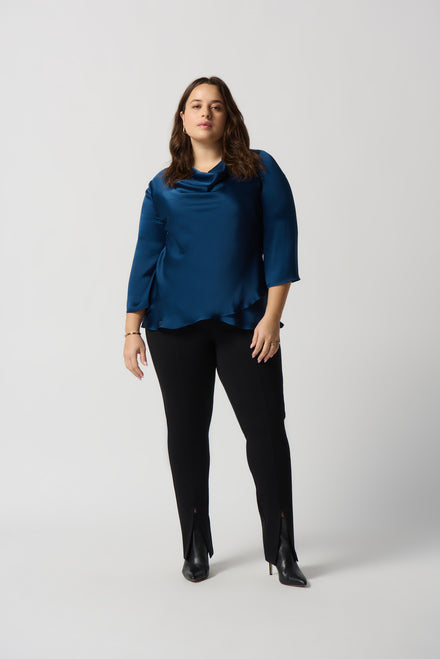 A person with short black hair is wearing a Royal Sapphire Draped Satin Blouse (Style 234082) from Joseph Ribkoff, featuring three-quarter sleeves and a tiered hem that adds an elegant touch. They are also wearing black pants. They are standing against a plain white background, gazing at the camera with arms loosely crossed.