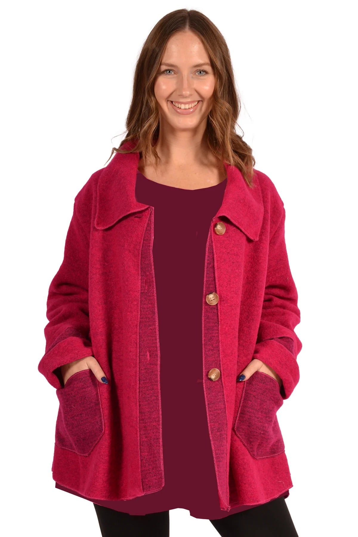 A smiling woman is wearing the Magenta Button Up Coat by Catherine Lillywhite, showcasing its exceptional quality with large buttons and two front pockets. Her hands rest in the pockets, and she has long, wavy hair. The background is plain white.