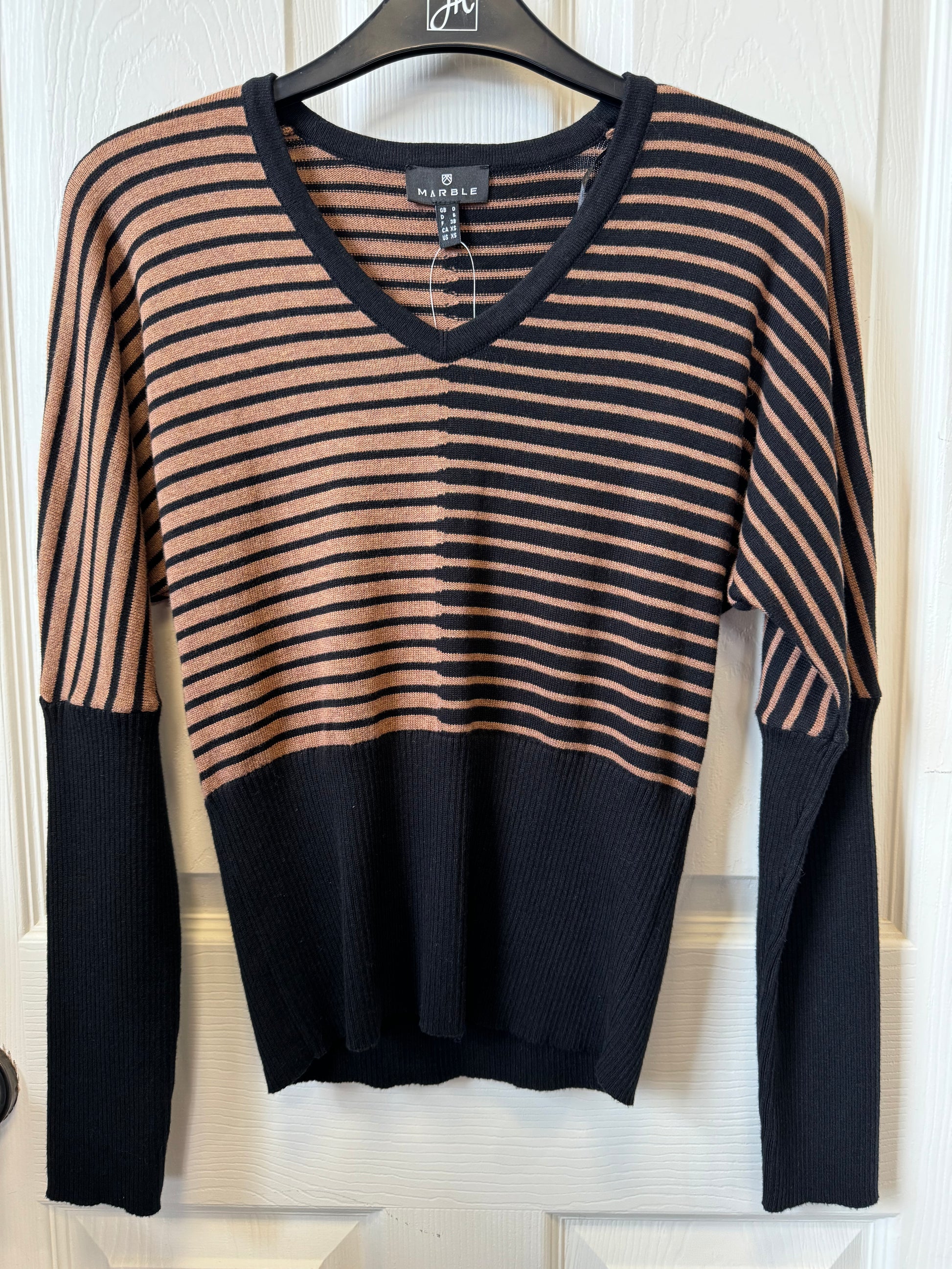 A Marble L/S Knit Tapered sweater by MARBLE hangs on a hanger against a white wooden background. The long-sleeved V-neck sweater features a black and tan stripe pattern on the top half and solid black from the chest down, including the sleeves' lower half.