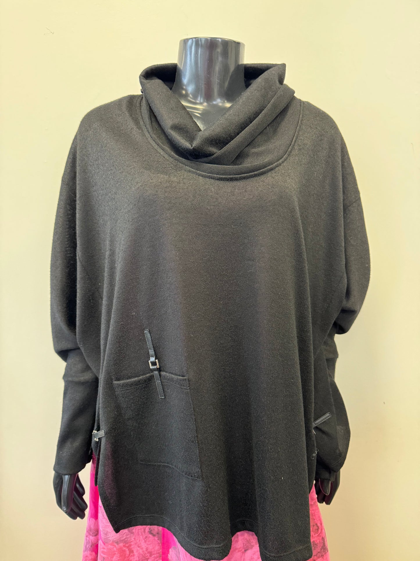 A black mannequin is dressed in a luxurious, loose-fitting, long-sleeve black sweater with a cowl neck and two front pockets. The sweater has subtle leather accents. The mannequin is also wearing a pink floral skirt with a vibrant pattern, making for an elegant Bali Throw Over - BLACK 7952 by Bali Corp ensemble.