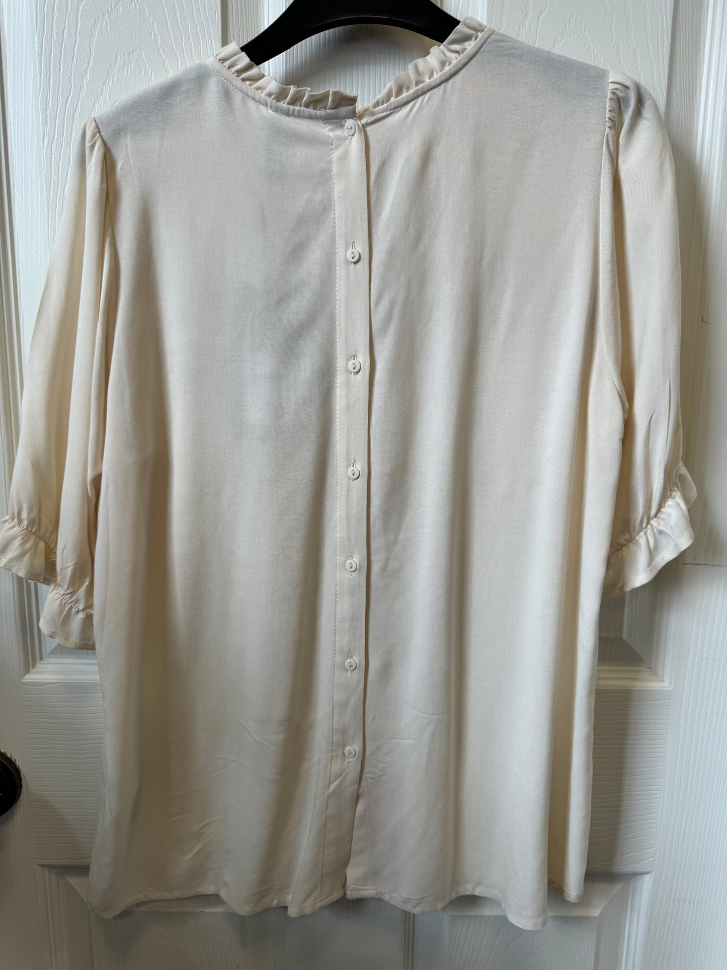 A Cream Button Up Shirt - SC-RADIA 128 from SoyaConcept, crafted from sustainable viscose, hangs on a black hanger against a white door. The blouse, designed like a button-up shirt, features a round neckline with ruffled trim, buttons running down the back, and short sleeves with ruffled cuffs.