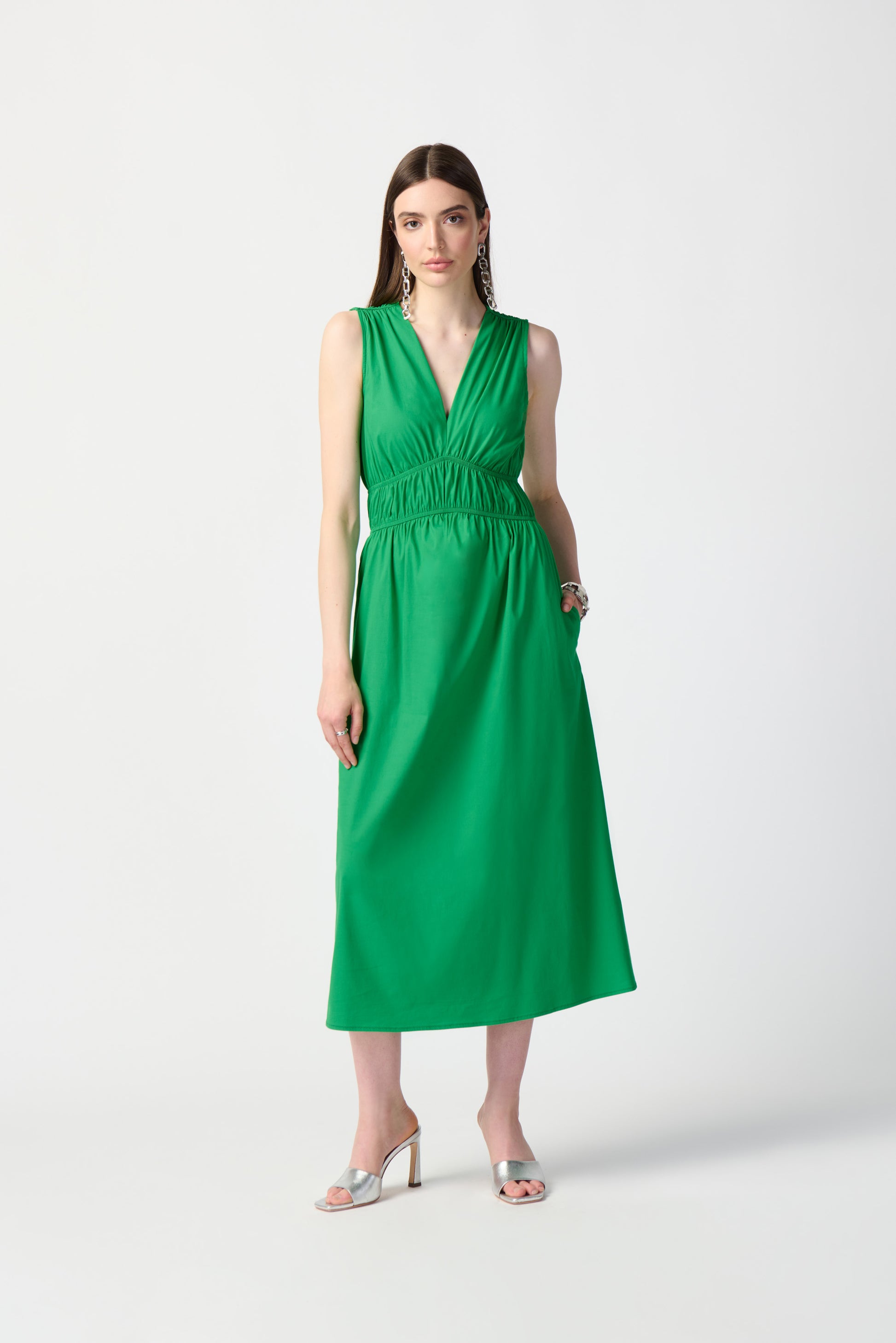 A woman stands against a plain, white background, wearing the Joseph Ribkoff Island Green Stretch Poplin Fit-and-Flare Dress 241127. This sleeveless, knee-length dress features woven fabric with gathered details at the waist. She has long, straight hair and is accessorized with drop earrings, bracelets, and open-toe high heels.