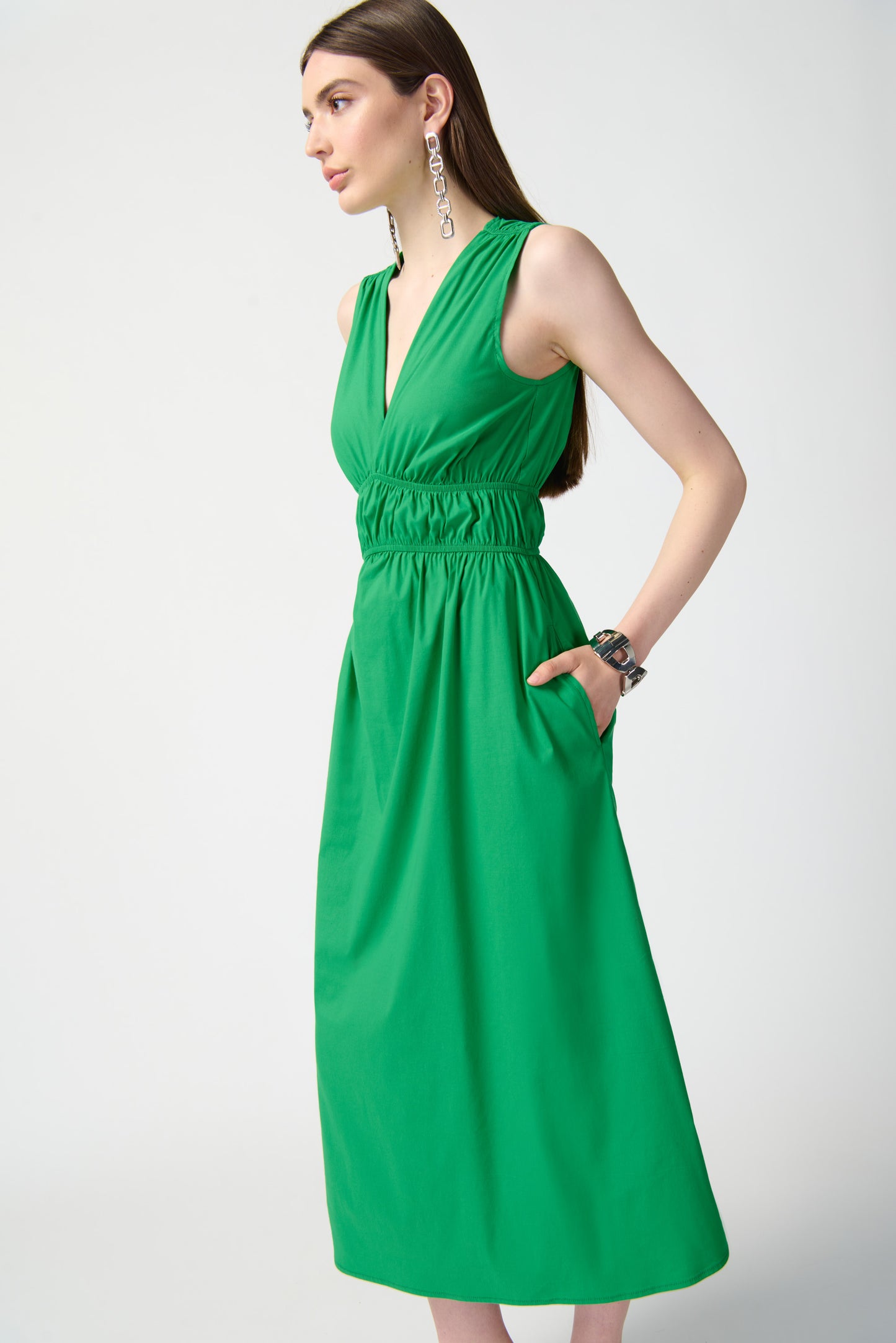 A woman stands against a plain, white background, wearing the Joseph Ribkoff Island Green Stretch Poplin Fit-and-Flare Dress 241127. This sleeveless, knee-length dress features woven fabric with gathered details at the waist. She has long, straight hair and is accessorized with drop earrings, bracelets, and open-toe high heels.