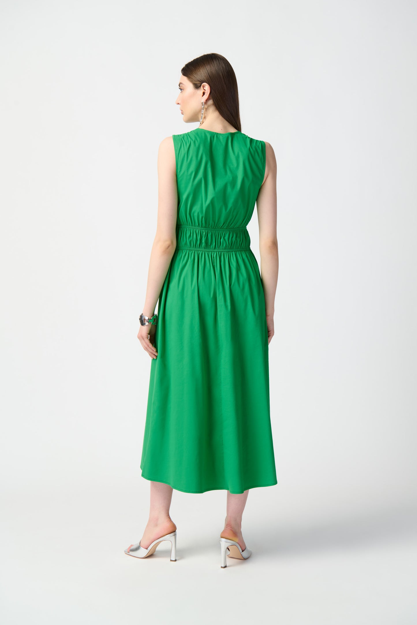 A woman stands against a plain, white background, wearing the Joseph Ribkoff Island Green Stretch Poplin Fit-and-Flare Dress 241127. This sleeveless, knee-length dress features woven fabric with gathered details at the waist. She has long, straight hair and is accessorized with drop earrings, bracelets, and open-toe high heels.