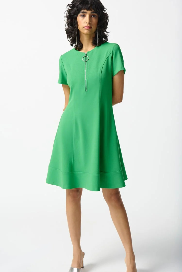 A person with shoulder-length dark wavy hair stands against a plain white background, wearing the Joseph Ribkoff - 242031 Island Green Scuba Crepe Fit and Flare Dress. The short-sleeved attire, featuring a zipped neckline and showcasing a fit and flare silhouette, is accessorized with a silver bracelet and white heeled sandals.