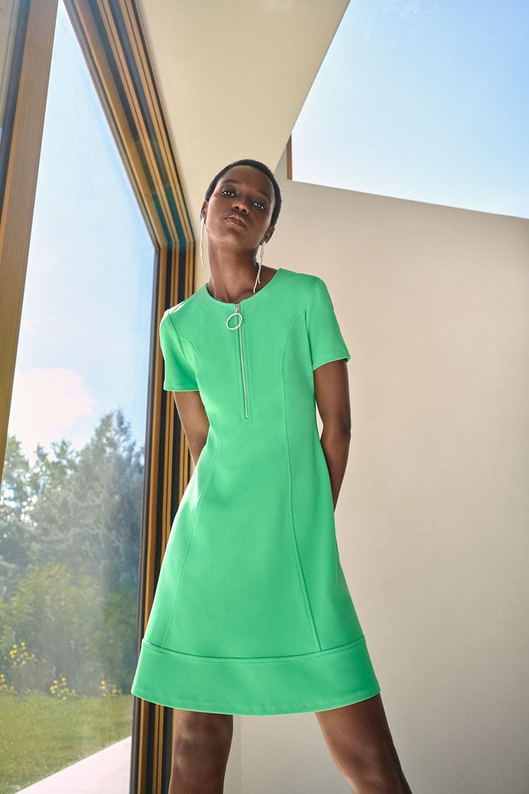 A person with shoulder-length dark wavy hair stands against a plain white background, wearing the Joseph Ribkoff - 242031 Island Green Scuba Crepe Fit and Flare Dress. The short-sleeved attire, featuring a zipped neckline and showcasing a fit and flare silhouette, is accessorized with a silver bracelet and white heeled sandals.