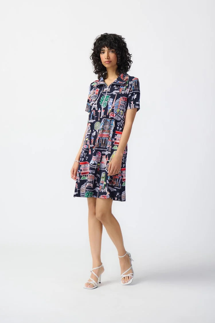 A person with dark, curly hair is wearing the Joseph Ribkoff - 241209 Scenery Print Silky Knit Trapeze Dress. Featuring a colorful cityscape pattern on a navy blue background, the short-sleeve dress has a zipped neckline and relaxed fit that falls above the knees. They are standing against a plain white backdrop, complemented by white strappy heels.