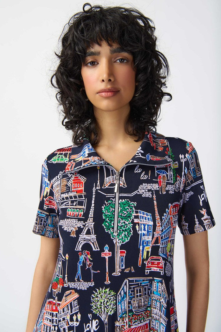 A person with dark, curly hair is wearing the Joseph Ribkoff - 241209 Scenery Print Silky Knit Trapeze Dress. Featuring a colorful cityscape pattern on a navy blue background, the short-sleeve dress has a zipped neckline and relaxed fit that falls above the knees. They are standing against a plain white backdrop, complemented by white strappy heels.