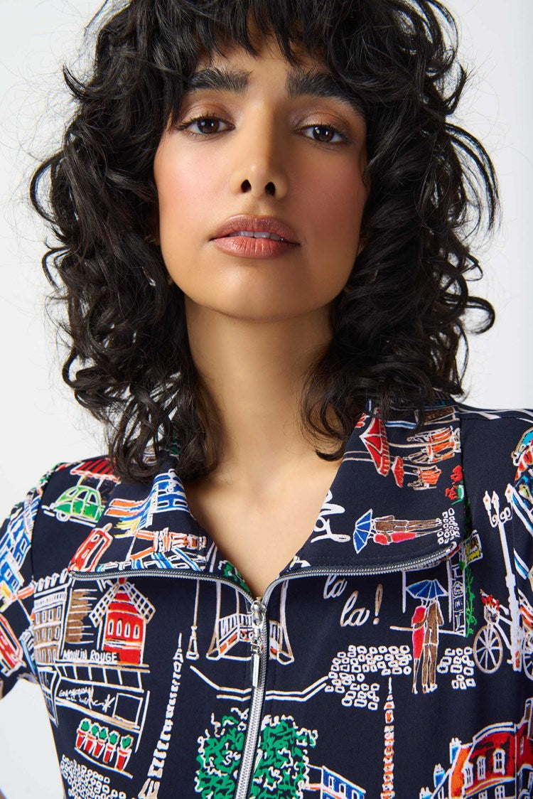 A person with dark, curly hair is wearing the Joseph Ribkoff - 241209 Scenery Print Silky Knit Trapeze Dress. Featuring a colorful cityscape pattern on a navy blue background, the short-sleeve dress has a zipped neckline and relaxed fit that falls above the knees. They are standing against a plain white backdrop, complemented by white strappy heels.