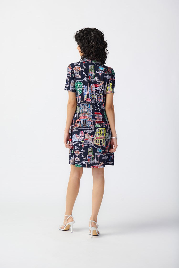 A person with dark, curly hair is wearing the Joseph Ribkoff - 241209 Scenery Print Silky Knit Trapeze Dress. Featuring a colorful cityscape pattern on a navy blue background, the short-sleeve dress has a zipped neckline and relaxed fit that falls above the knees. They are standing against a plain white backdrop, complemented by white strappy heels.