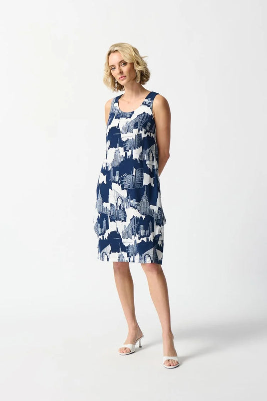 A person with short, blonde hair stands against a white background. They are wearing the Joseph Ribkoff - Gauze Scenery-Print Cocoon Dress 242157, a sleeveless, knee-length dress featuring pleat details and an abstract blue and white pattern, complemented by white high-heeled sandals. The dress has a back zipper opening and their hands are behind their back.