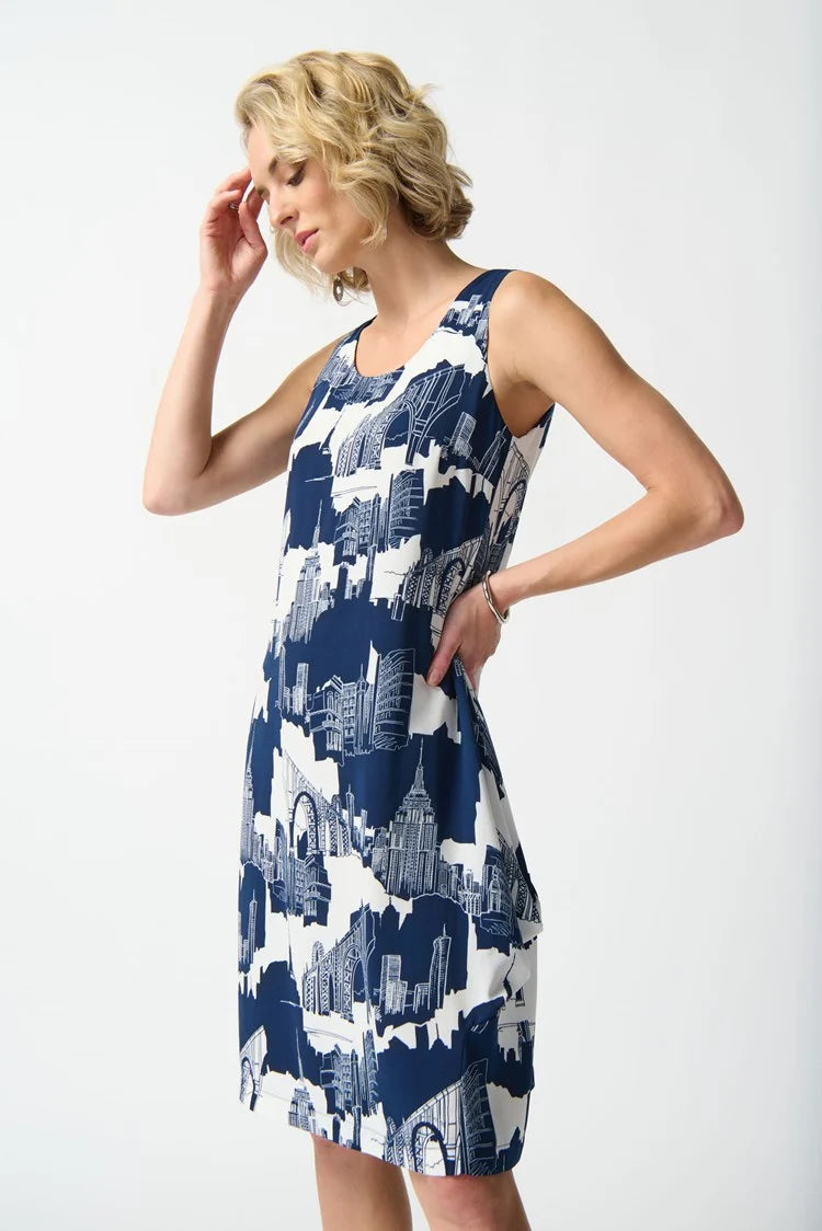 A person with short, blonde hair stands against a white background. They are wearing the Joseph Ribkoff - Gauze Scenery-Print Cocoon Dress 242157, a sleeveless, knee-length dress featuring pleat details and an abstract blue and white pattern, complemented by white high-heeled sandals. The dress has a back zipper opening and their hands are behind their back.