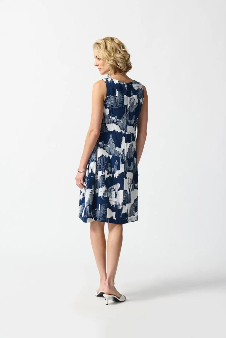 A person with short, blonde hair stands against a white background. They are wearing the Joseph Ribkoff - Gauze Scenery-Print Cocoon Dress 242157, a sleeveless, knee-length dress featuring pleat details and an abstract blue and white pattern, complemented by white high-heeled sandals. The dress has a back zipper opening and their hands are behind their back.