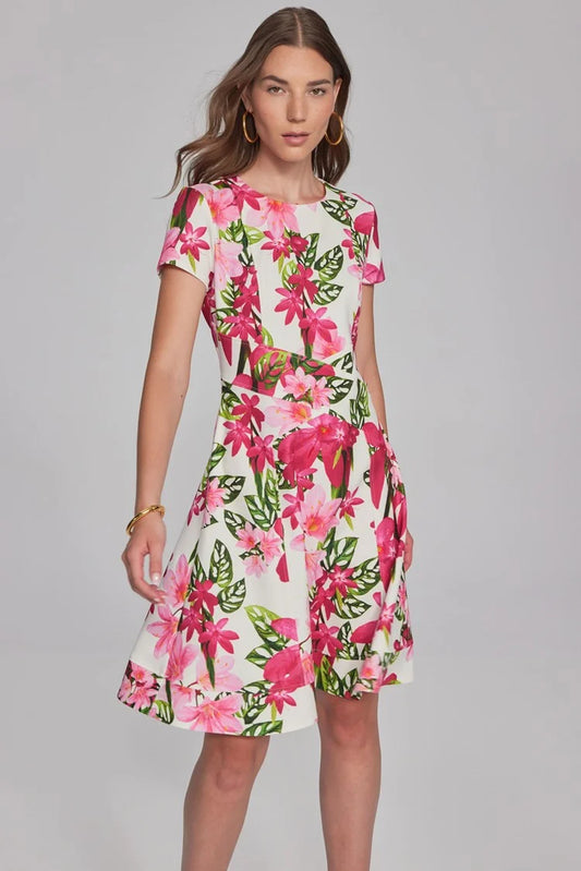 A woman with long brown hair, wearing hoop earrings and a bracelet, is dressed in the Joseph Ribkoff Floral Print Scuba Crepe Fit-And-Flare Dress 241789. This short-sleeved, knee-length white dress features vibrant pink and green floral patterns from the renowned brand Joseph Ribkoff. The fit-and-flare silhouette ensemble beautifully complements her as she stands against a plain, light gray background.