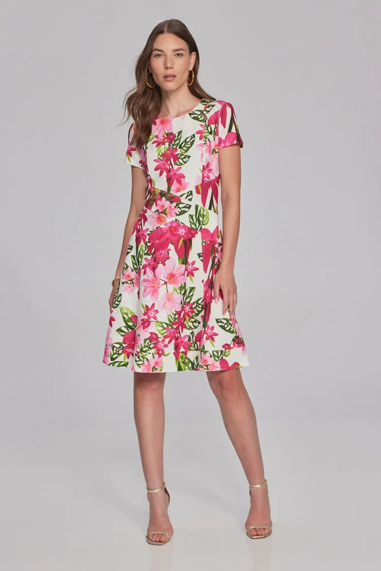 A woman with long brown hair, wearing hoop earrings and a bracelet, is dressed in the Joseph Ribkoff Floral Print Scuba Crepe Fit-And-Flare Dress 241789. This short-sleeved, knee-length white dress features vibrant pink and green floral patterns from the renowned brand Joseph Ribkoff. The fit-and-flare silhouette ensemble beautifully complements her as she stands against a plain, light gray background.