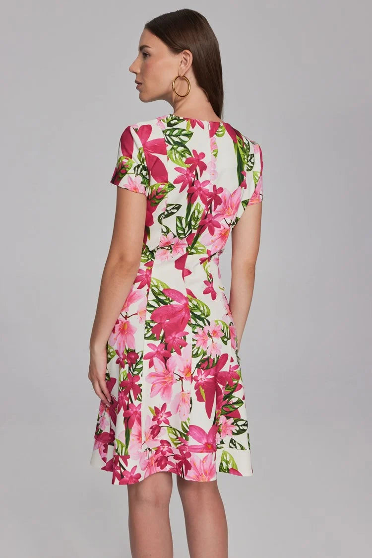 A woman with long brown hair, wearing hoop earrings and a bracelet, is dressed in the Joseph Ribkoff Floral Print Scuba Crepe Fit-And-Flare Dress 241789. This short-sleeved, knee-length white dress features vibrant pink and green floral patterns from the renowned brand Joseph Ribkoff. The fit-and-flare silhouette ensemble beautifully complements her as she stands against a plain, light gray background.