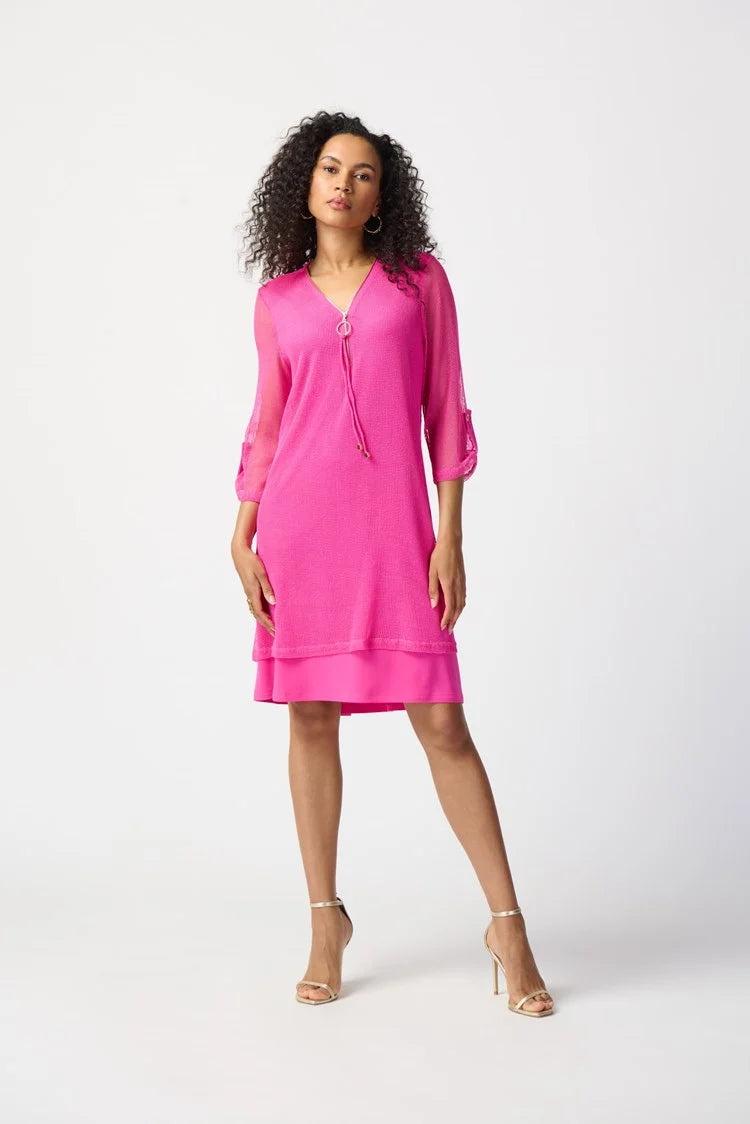 A woman with curly hair, wearing the Joseph Ribkoff - Bouclé Mesh and Silky Knit Layered Dress 241115 in a vibrant pink hue, poses confidently against a plain white background. The knee-length dress features a zipped neckline and falls just above her knees. With one hand resting on her hip, she completes the look with strappy high-heeled sandals.