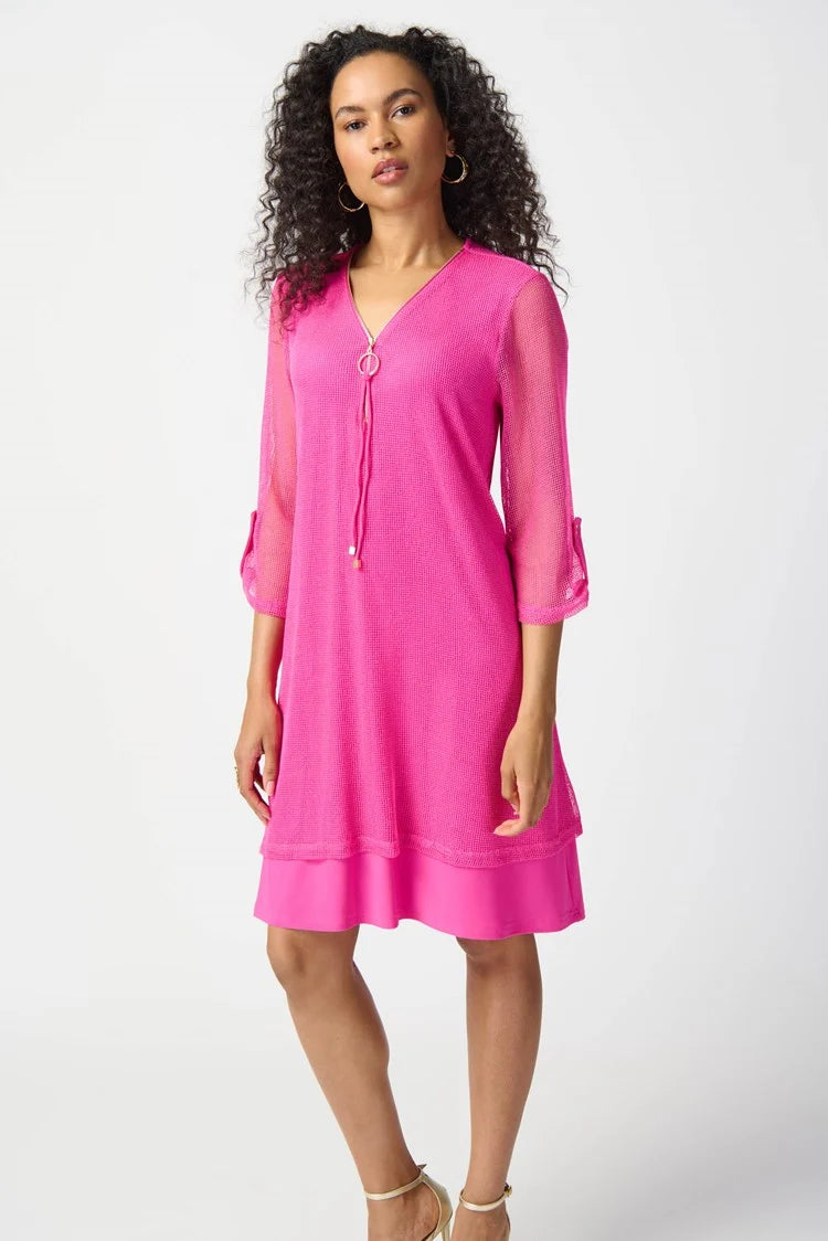 A woman with curly hair, wearing the Joseph Ribkoff - Bouclé Mesh and Silky Knit Layered Dress 241115 in a vibrant pink hue, poses confidently against a plain white background. The knee-length dress features a zipped neckline and falls just above her knees. With one hand resting on her hip, she completes the look with strappy high-heeled sandals.