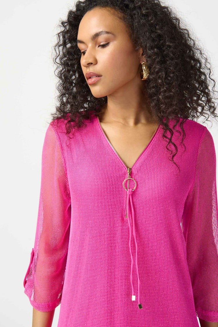 A woman with curly hair, wearing the Joseph Ribkoff - Bouclé Mesh and Silky Knit Layered Dress 241115 in a vibrant pink hue, poses confidently against a plain white background. The knee-length dress features a zipped neckline and falls just above her knees. With one hand resting on her hip, she completes the look with strappy high-heeled sandals.