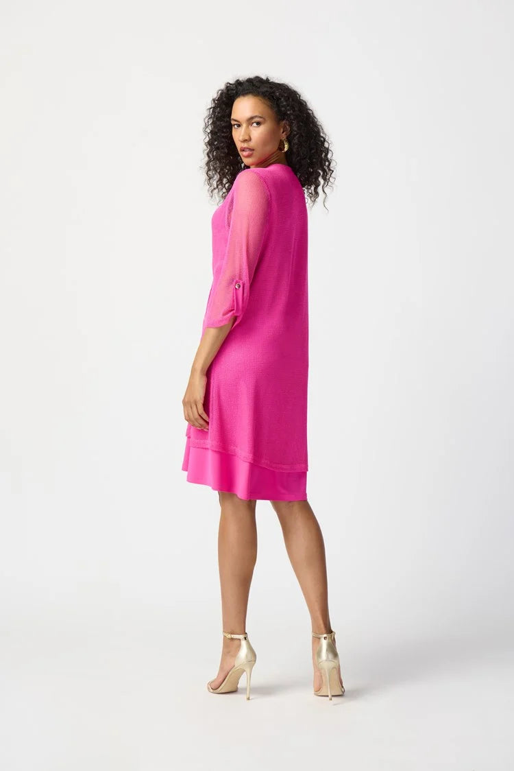 A woman with curly hair, wearing the Joseph Ribkoff - Bouclé Mesh and Silky Knit Layered Dress 241115 in a vibrant pink hue, poses confidently against a plain white background. The knee-length dress features a zipped neckline and falls just above her knees. With one hand resting on her hip, she completes the look with strappy high-heeled sandals.