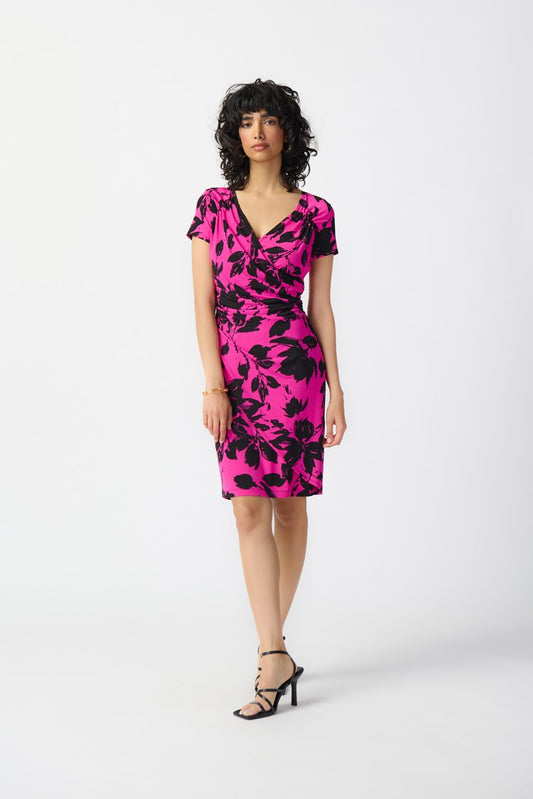 A woman with curly black hair is modeling the Joseph Ribkoff - Floral Print Silky Knit Wrap Dress 241118. The pink dress, featuring a black floral pattern, showcases a contemporary style with its knee-length hemline, short sleeves, and v-neckline. She pairs it with black strappy high-heeled sandals and stands against a plain white background.