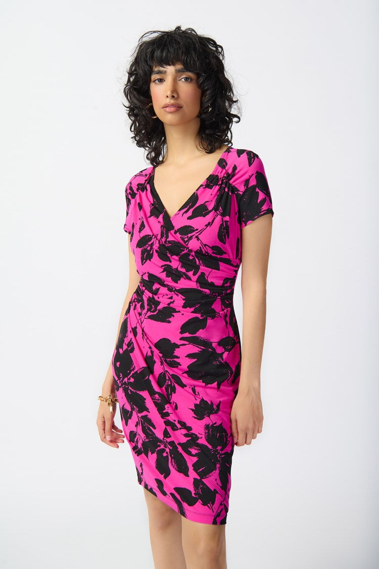 A woman with curly black hair is modeling the Joseph Ribkoff - Floral Print Silky Knit Wrap Dress 241118. The pink dress, featuring a black floral pattern, showcases a contemporary style with its knee-length hemline, short sleeves, and v-neckline. She pairs it with black strappy high-heeled sandals and stands against a plain white background.