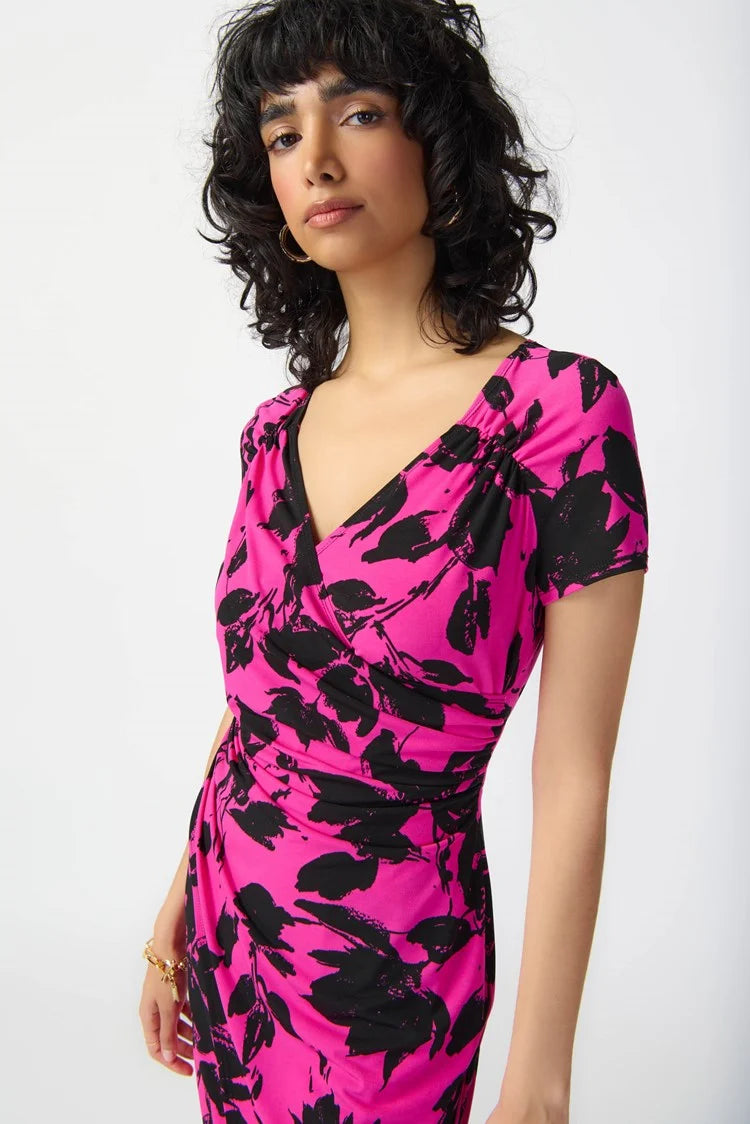 A woman with curly black hair is modeling the Joseph Ribkoff - Floral Print Silky Knit Wrap Dress 241118. The pink dress, featuring a black floral pattern, showcases a contemporary style with its knee-length hemline, short sleeves, and v-neckline. She pairs it with black strappy high-heeled sandals and stands against a plain white background.