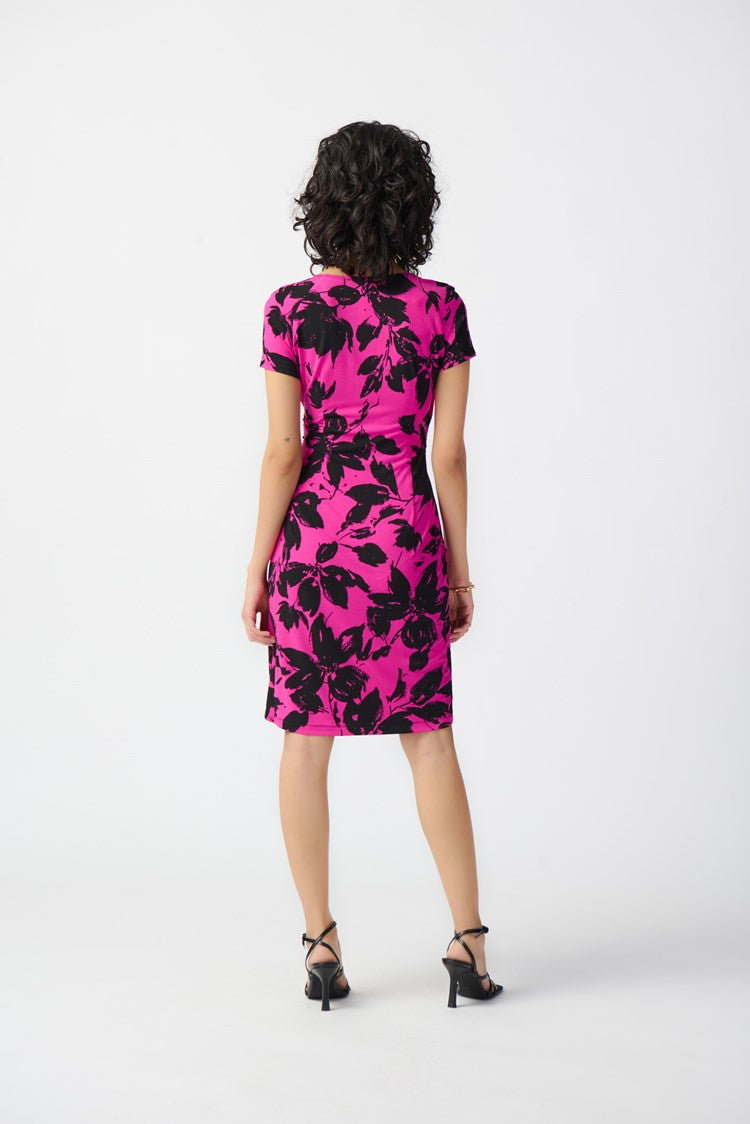 A woman with curly black hair is modeling the Joseph Ribkoff - Floral Print Silky Knit Wrap Dress 241118. The pink dress, featuring a black floral pattern, showcases a contemporary style with its knee-length hemline, short sleeves, and v-neckline. She pairs it with black strappy high-heeled sandals and stands against a plain white background.