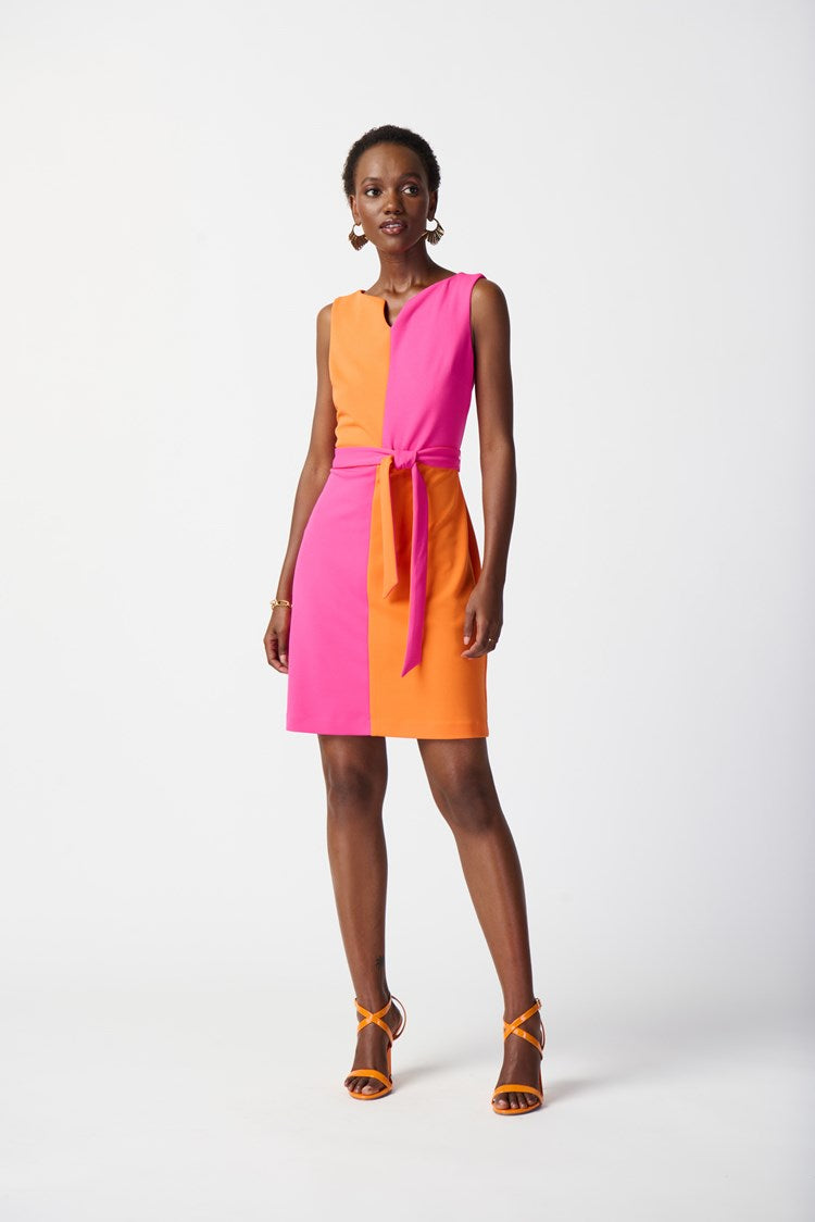 A woman stands confidently modeling the Joseph Ribkoff Scuba Crepe Colour-Block Shift Dress 241193. The sleeveless dress, crafted from scuba crepe fabric, features a striking colour-block pattern with one half in bright pink and the other in orange. A matching pink belt is tied at the waist to accentuate her figure. She completes her look with orange high-heeled sandals and gold jewelry.
