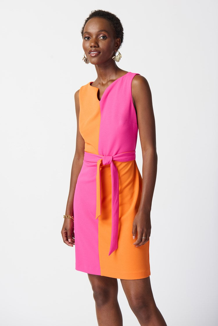 A woman stands confidently modeling the Joseph Ribkoff Scuba Crepe Colour-Block Shift Dress 241193. The sleeveless dress, crafted from scuba crepe fabric, features a striking colour-block pattern with one half in bright pink and the other in orange. A matching pink belt is tied at the waist to accentuate her figure. She completes her look with orange high-heeled sandals and gold jewelry.
