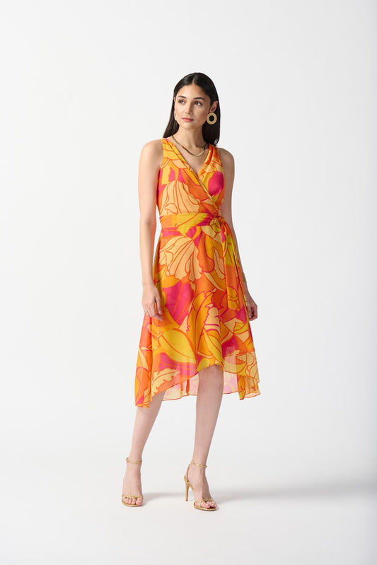 A woman stands against a plain white background, wearing the Joseph Ribkoff Chiffon Tropical Print Fit and Flare Dress 242015. The sleeveless, knee-length dress features a bold, colorful floral pattern in shades of orange, pink, and yellow. She pairs it with nude high-heeled sandals and large hoop earrings.