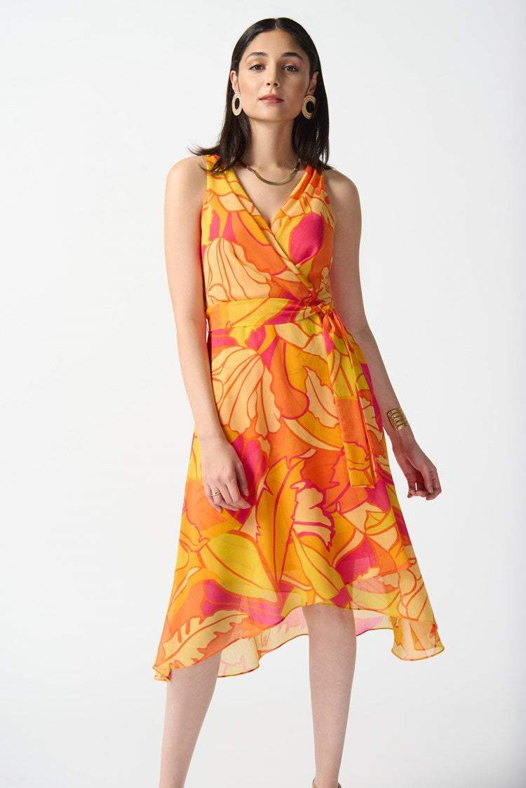 A woman stands against a plain white background, wearing the Joseph Ribkoff Chiffon Tropical Print Fit and Flare Dress 242015. The sleeveless, knee-length dress features a bold, colorful floral pattern in shades of orange, pink, and yellow. She pairs it with nude high-heeled sandals and large hoop earrings.