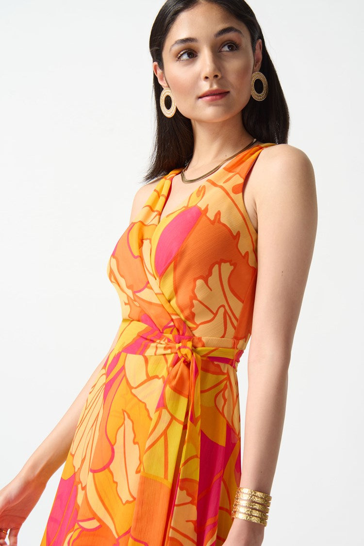 A woman stands against a plain white background, wearing the Joseph Ribkoff Chiffon Tropical Print Fit and Flare Dress 242015. The sleeveless, knee-length dress features a bold, colorful floral pattern in shades of orange, pink, and yellow. She pairs it with nude high-heeled sandals and large hoop earrings.