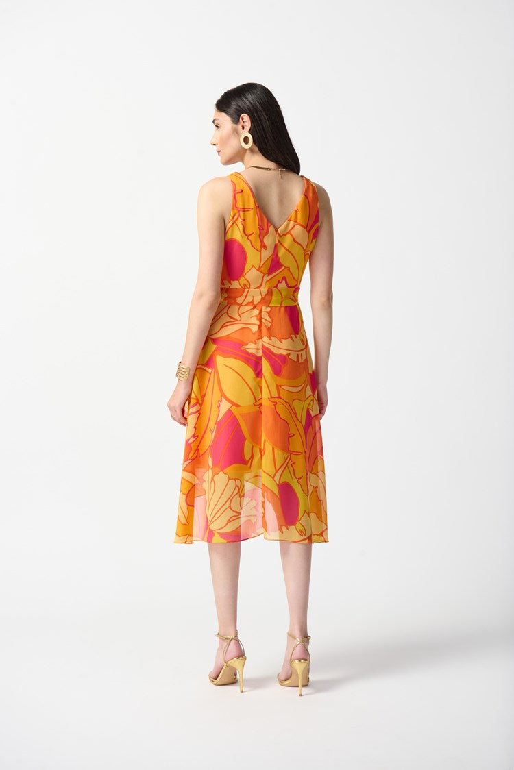 A woman stands against a plain white background, wearing the Joseph Ribkoff Chiffon Tropical Print Fit and Flare Dress 242015. The sleeveless, knee-length dress features a bold, colorful floral pattern in shades of orange, pink, and yellow. She pairs it with nude high-heeled sandals and large hoop earrings.