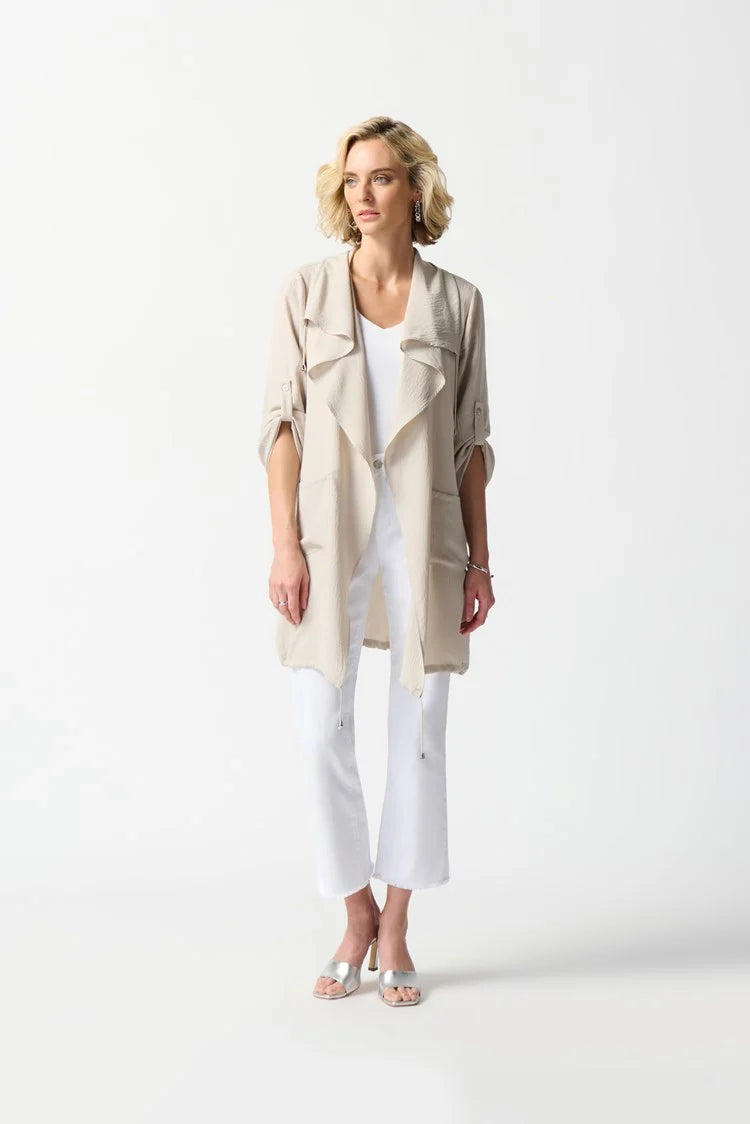 A person stands against a plain white background wearing the Joseph Ribkoff Gauze Boxy Cover Up in Moonstone with a shawl collar over a white top, white pants, and white heeled sandals. The individual has short blonde hair styled in loose waves and poses with their head turned slightly to the right.